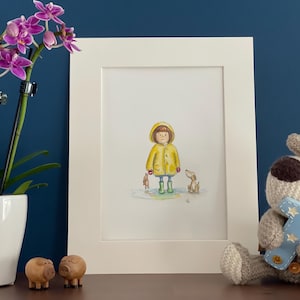 Puede incluir: Una impresión en acuarela enmarcada representa a un niño con impermeable amarillo y botas verdes, de pie en un charco con un perro. Un pequeño peluche y figuras de cerdos de madera están en el estante. Una orquídea blanca en una maceta blanca está a la izquierda.