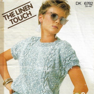 Pode incluir: Um top de malha azul claro com um design de malha trançada. O top tem mangas curtas e um decote redondo. A modelo está usando o top com um par de shorts jeans azul claro. O texto "THE LINEN TOUCH" está impresso na parte superior da imagem. O texto "DK 6762 30-40" está impresso no canto superior direito da imagem. O texto "Sirdar Sunseeker" está impresso no canto inferior direito da imagem.