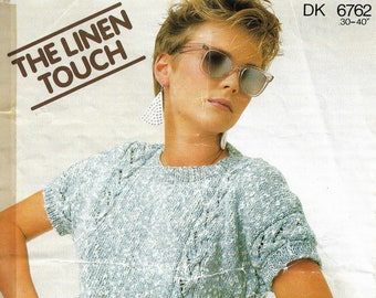May include: A light blue knitted top with a cable knit design. The top has short sleeves and a round neckline. The model is wearing the top with a pair of light blue denim shorts. The text "THE LINEN TOUCH" is printed on the top of the image. The text "DK 6762 30-40" is printed in the top right corner of the image. The text "Sirdar Sunseeker" is printed in the bottom right corner of the image.