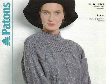 May include: A gray cable knit sweater with a high neck and long sleeves. The sweater is made with a speckled yarn and has a relaxed fit. The model is wearing a black hat and a black skirt.