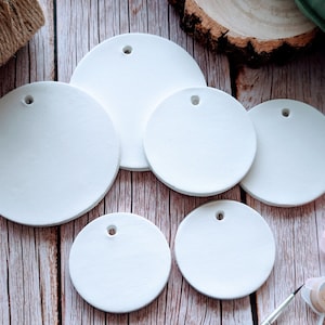 May include: Five white ceramic round ornaments with holes for hanging. The ornaments are on a wooden surface with paint supplies.