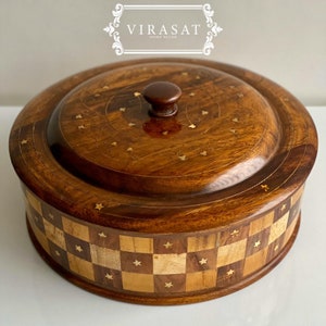 Puede incluir: Un plato de servir redondo de madera con tapa. El plato está hecho de madera oscura con un patrón de tablero de ajedrez y estrellas doradas. La tapa tiene un diseño decorativo con acentos dorados. El plato es perfecto para servir aperitivos, salsas o postres.