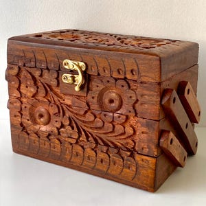 May include: A rectangular wooden box with detailed floral carvings and a brass clasp. The box is stained a warm brown colour and features relief patterns on the lid and sides. The box has a hinged design with wooden supports.