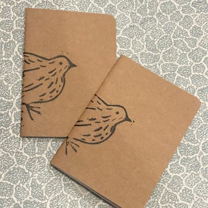 Peut inclure: Deux carnets marrons avec une simple illustration d'oiseau noir sur la couverture. Les carnets sont sur une surface à motifs.