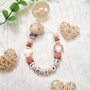 Détail d’une attache tétine personnalisée arc en ciel et coeur – combinaison de perles terracotta et clip bois naturel – fait main en France.