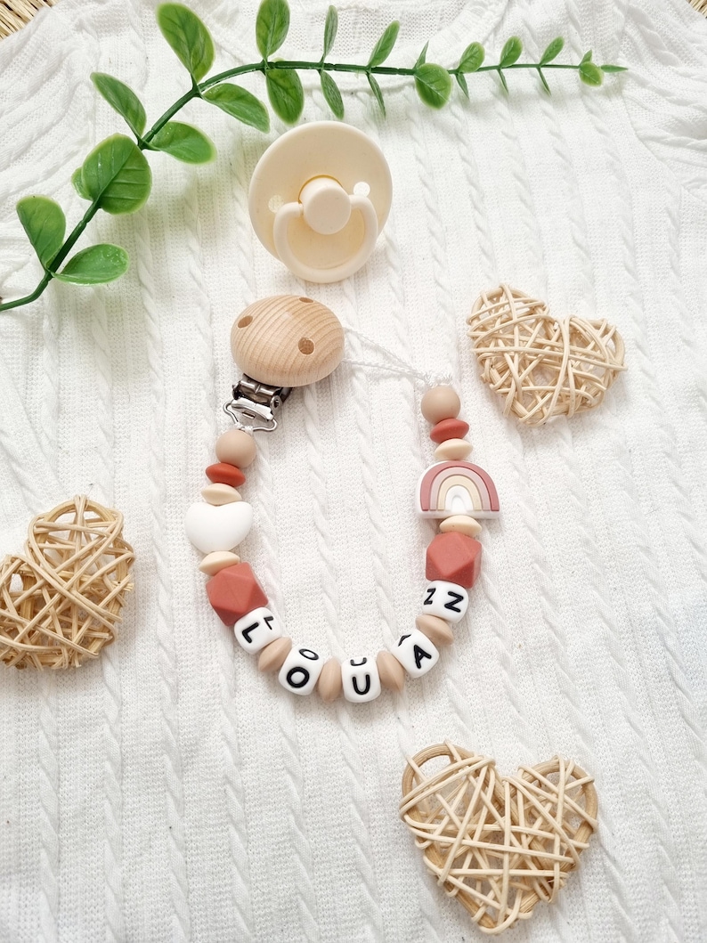 Détail d’une attache tétine personnalisée arc en ciel et coeur – combinaison de perles terracotta et clip bois naturel – fait main en France.