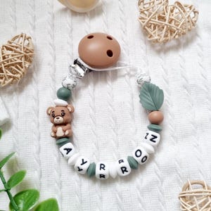 Peut inclure: Un attache-tétine personnalisé pour bébé avec le nom "AYROR" épelé en perles de lettres blanches. Le clip comprend un charm en forme d'ours brun, des perles vertes et blanches et un support de tétine marron.