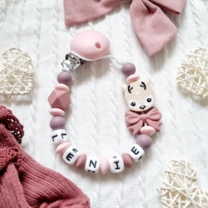 Może przedstawiać: Różowy i fioletowy klips do smoczka dla dziecka z imieniem "LENNIE" zapisanym białymi koralikami z literami. Klips posiada zawieszkę w kształcie jelenia z kokardą i różowy smoczek. Tło to biała, teksturowana powierzchnia.