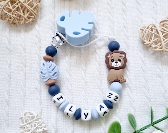 May include: A pacifier clip featuring a light blue silicone monstera leaf clip, a brown lion-shaped bead, and round beads in shades of navy blue, light blue, and brown. White cube beads spell out the name ILYANZ in black letters on the string.