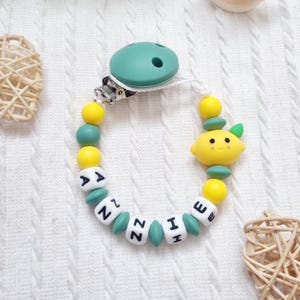 Può includere: Una catenella portaciuccio con una clip in silicone verde acqua attaccata a una serie di perline. Le perline si alternano tra sfere gialle e verde acqua e cubi bianchi con lettere nere che compongono ANNIE. È inclusa una perlina a forma di limone.