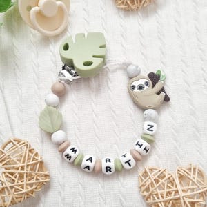 May include: A personalized pacifier clip featuring a silicone sloth charm, a sage green monstera leaf clip, and white beads spelling the name MARTIN. The design includes round beige, white, and marbled silicone beads strung on a white cord.