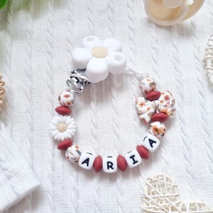 May include: A baby pacifier clip with the name "ARIA" spelled out in white letter beads. The clip features white flower and floral patterned beads, and maroon-coloured beads. A white flower-shaped clip is at the top.
