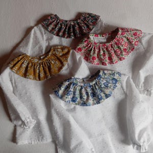 Peut inclure: Quatre chemises blanches à manches longues en tissu plumetis, chacune dotée d'un col à volants à motifs floraux. Les cols présentent des palettes de couleurs variées, notamment des imprimés floraux bleu foncé, jaune moutarde, rose et bleu clair.