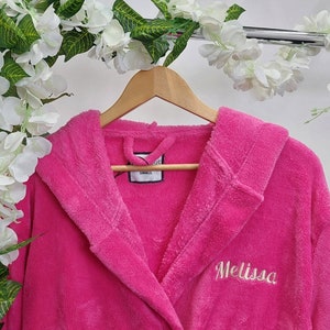 May include: A bright pink, hooded bathrobe with the name "Melissa" embroidered in a cursive font. The robe is hanging on a wooden hanger. A tag reads "SMALL". The background includes white flowers and greenery.