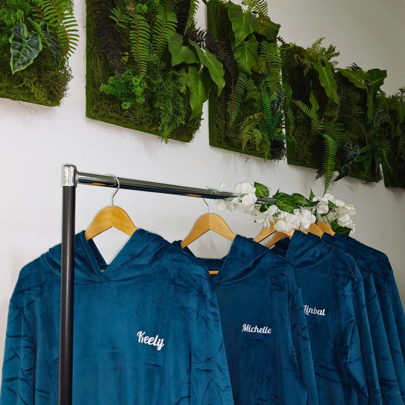 May include: Four teal blue hooded sweatshirts with white embroidered names: Keely, Michelle, Linbat, and another name. The sweatshirts are hanging on a rack with wooden hangers.