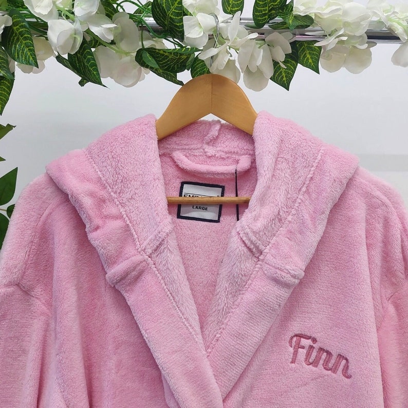 May include: A light pink plush bathrobe with a hood, hanging on a wooden hanger. The word "Finn" is embroidered on the front in a matching pink thread. A tag reads "LARGE".