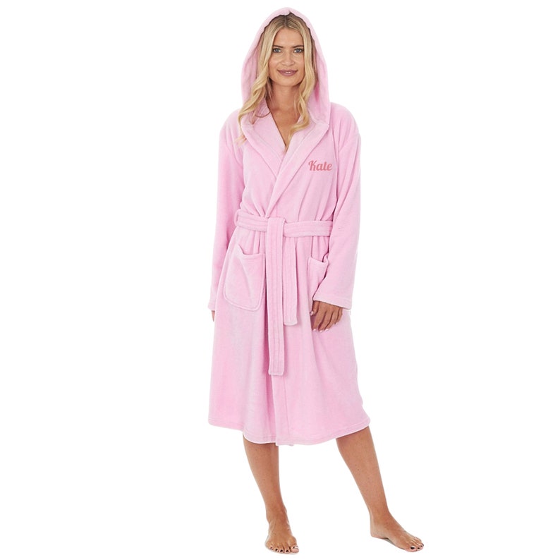 May include: A light pink hooded bathrobe with a matching belt and pockets. The word "Kate" is embroidered on the chest. The robe is made of a soft, plush material.