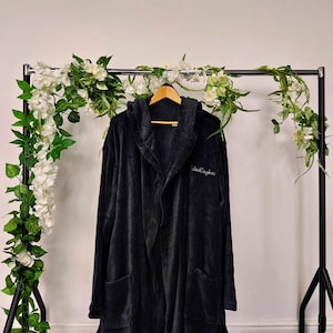 May include: A dark grey hooded robe with pockets hangs on a wooden hanger. The robe has the embroidered text "Jack Caybor" on the front. The robe is displayed on a black metal clothing rack decorated with white flowers and greenery.