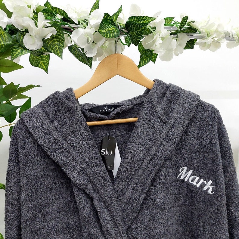 May include: Gray hooded robe with a white embroidered name "Mark" on the front. The robe is hanging on a wooden hanger.