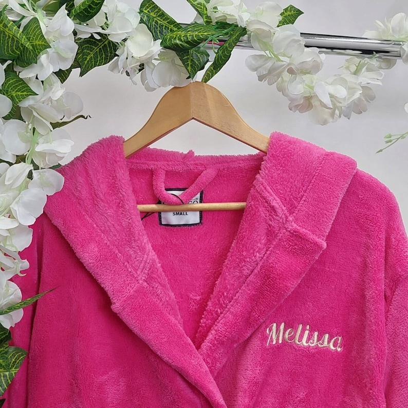 May include: A bright pink, hooded bathrobe with the name "Melissa" embroidered in a cursive font. The robe is hanging on a wooden hanger. A tag reads "SMALL". The background includes white flowers and greenery.