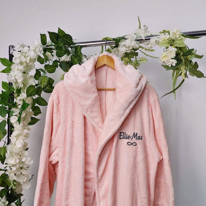May include: A light pink plush robe with a hood, hanging on a wooden hanger. The robe has the name "Ellie Mac" embroidered in dark blue, along with an infinity symbol. The robe is displayed on a metal rack with floral decorations.