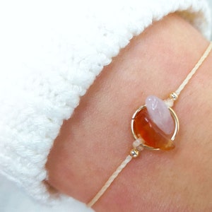 May include: A delicate bracelet featuring a gold ring holding a carnelian and rose quartz stone. The bracelet is made with a thin white cord.