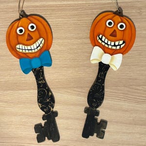 May include: Two hanging ornaments shaped like keys with orange pumpkin heads featuring wide grins and large eyes. One pumpkin wears a bright blue bow tie, while the other wears a cream-colored bow tie. The key shafts are black with a swirling pattern.