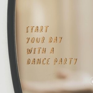 Könnte beinhalten: Ein runder goldfarbener Spiegel mit schwarzem Rahmen. Der Spiegel hat den Text "Start your day with a dance party" in die Oberfläche graviert.