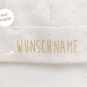 Könnte beinhalten: Weißer Babyhut mit rosa Punkten und goldener Aufschrift "Wunschname". Der Hut hat ein Etikett mit der Aufschrift "aufbügler".