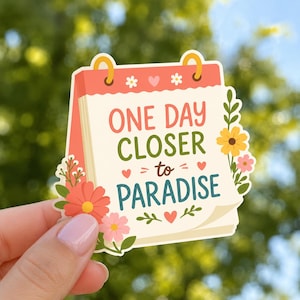 May include: A sticker in the shape of a calendar with the words "ONE DAY CLOSER to PARADISE" in coral, green, and teal. The sticker has floral and heart accents, with a peach-colored top and gold loops.