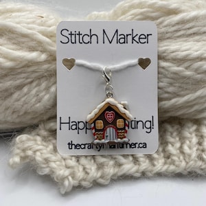Op de afbeelding: Een gingerbread house steekmarkeerder met een wit dak en rood hartdetail hangt aan een witte kaart met de tekst "Stitch Marker" en "Happy Stitching!". De markeerder is bevestigd aan een witte draad en rust op crèmekleurig garen.