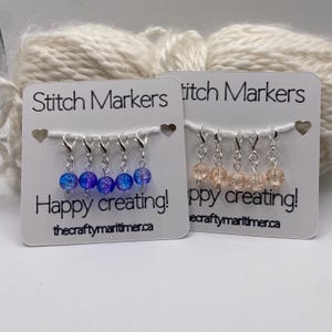 May include: Two white display cards labeled Stitch Markers and Happy creating! with thecraftymaritimer.ca printed at the bottom. Each card holds five silver-toned lobster clasp stitch markers with round crackle glass beads in blue and peach.