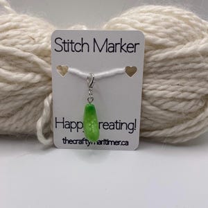 May include: A green pickle-shaped stitch marker with a silver clasp hangs from a white card that reads "Stitch Marker" and "Happy creating!" The background features a ball of off-white yarn.
