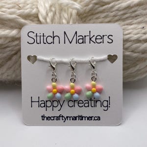 May include: A white card with the text "Stitch Markers" and "Happy creating!" displays three flower-shaped stitch markers. The markers are pink, yellow, blue, and green, attached to silver clasps. The website address thecraftymaritime.ca is also visible.