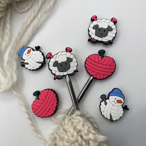 May include: Knitting needle accessories with sheep, snowmen, and yarn ball designs in white, black, red, and blue. The silver needles are inserted into a knitted piece, showcasing a crafting theme.