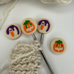 May include: Two metal knitting needles with round, cream-coloured end caps featuring orange jack-o'-lanterns and purple ghost designs. Two additional loose round caps with the same pumpkin and ghost motifs are placed nearby on a white surface.