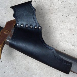 Puede incluir: Una vaina de cuero negro con una parte superior en forma de murciélago decorativa y tachuelas plateadas. La vaina está diseñada para sostener una espada o una daga.