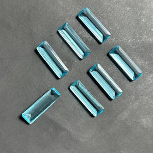 Puede incluir: Seis piedras preciosas rectangulares de color azul claro con facetas dispuestas en un patrón disperso. Cada piedra presenta una calidad transparente y clara con bordes afilados y una superficie pulida que refleja la luz en su forma alargada.