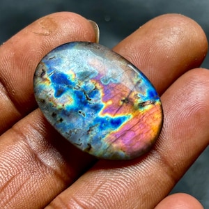 May include: An oval-shaped labradorite gemstone featuring a polished surface with iridescent flashes of blue, purple, and orange colors. The stone displays natural internal fractures and a smooth, rounded edge throughout its entire perimeter.