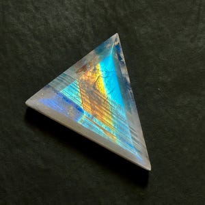 May include: A triangular-shaped gemstone with a translucent, clear appearance. The interior displays a vibrant play of colour, featuring distinct bands of iridescent blue and warm golden-yellow light that create a glowing, multi-faceted effect.