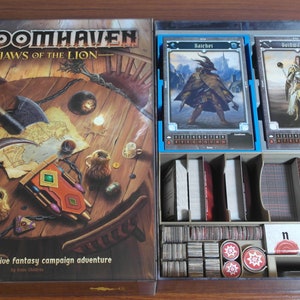 May include: A board game box with a brown wooden table and various objects on it. The box has the title "Gloomhaven: Jaws of the Lion" and the text "A fully cooperative fantasy campaign adventure". The box is open and shows a variety of game components, including cards, tokens, and dice.