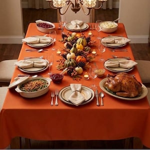 May include: A dining table set with an orange tablecloth, featuring a roasted turkey, serving dishes with green beans and cranberry sauce, plates with cream-colored napkins, wine glasses, silverware, and a centerpiece of small pumpkins and pinecones.