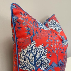May include: A decorative pillow with a vibrant red background and a blue and white Asian-inspired tree and landscape design. The pillow has a blue trim and is a square shape.