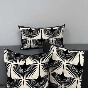 May include: Decorative throw pillows with a black and white crane pattern. The pillows have a black border and a cream-colored background. The crane design is repeated across the surface of the pillows. The pillows are arranged on a gray sofa.