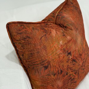 May include: Square decorative pillow with a textured, rust-colored fabric. The pillow has a detailed pattern with shades of brown and orange. The pillow is resting on a white surface.