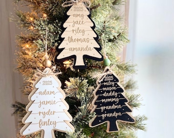 May include: Three wooden Christmas tree ornaments with a black, white, and natural wood finish. Each ornament features a year, 2023, and a list of names in gold lettering. The names on the black ornament are Miley, Mason, Daddy, Mommy, Grandma, and Grandpa. The names on the white ornament are Adam, Jamie, Ryder, and Finley. The names on the natural wood ornament are Amy, Jace, Riley, Thomas, and Amanda.