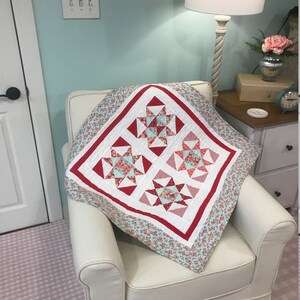 May include: A red and white quilt with a floral pattern. The quilt is made up of four squares, each with a different design. The quilt is sitting on a white armchair.