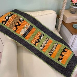 May include: A Halloween-themed table runner with a patchwork design. The runner features orange, black, and green fabrics with pumpkin and bat patterns. The runner is bordered with a dark gray fabric and green trim.