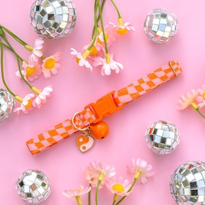 May include: Orange and pink checkered cat collar with a matching orange buckle and bell. The collar has a gold-colored ring with a heart-shaped charm featuring a yin-yang design. The background is pink with disco balls and flowers.