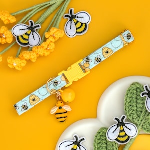 May include: A light blue and white patterned dog collar with a bee design and a gold bell charm. The collar is on a white and green crocheted surface.